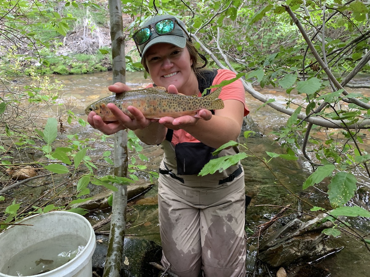 ISU student by stream holding up a rainbow trout.