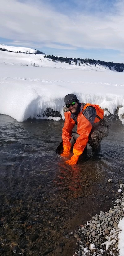 Jeremy Brooks in the middle of a stream collecting insects in the winter, with the banks covered in snow. 