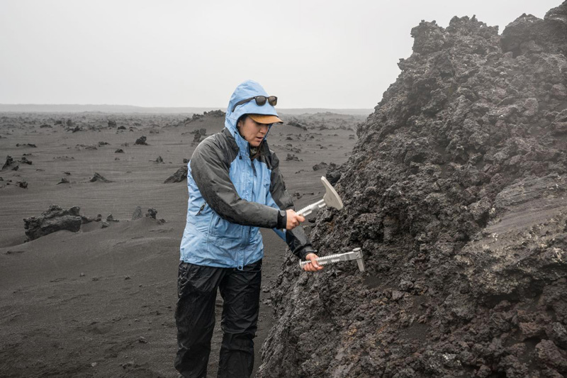 Angela Garcia demonstrates how to use a rock hammer and chisel to dislodge a rock sample from a large boulder during the Artemis II field geology training in Iceland. Credit: NASA